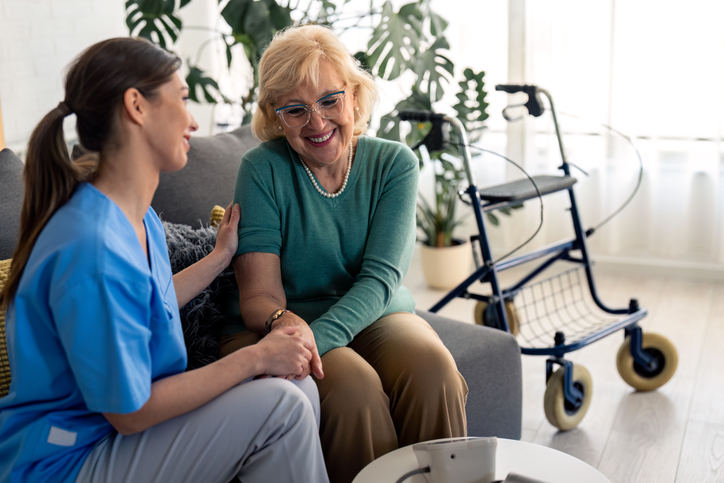 Female home care specialist and satisfied elderly woman holding hands while talking and sitting in living room.